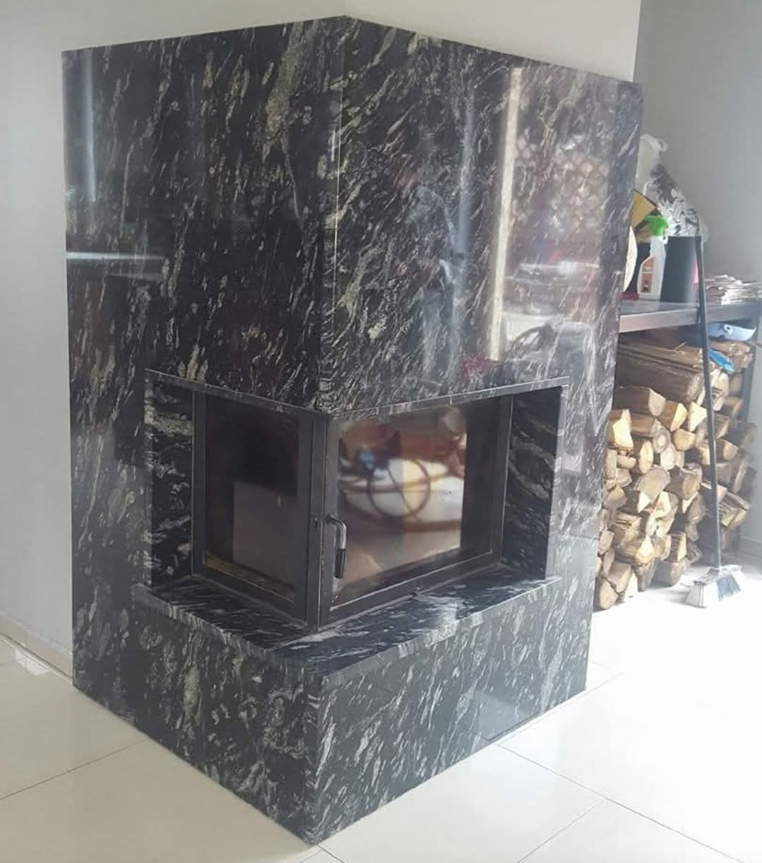 Obudowa kominka z ciemnego kamienia z wyraźnym wzorem, metalowe drzwiczki. W tle widoczne drewno na półce, jasna podłoga.