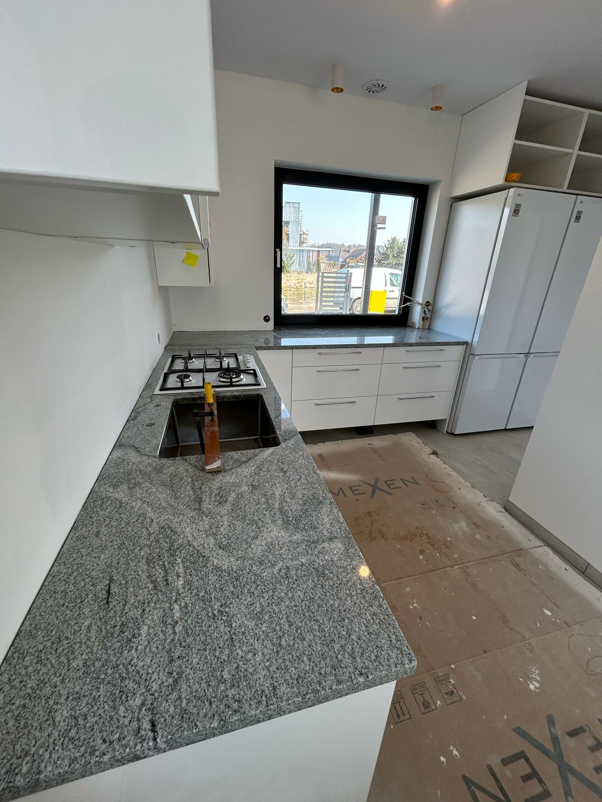 Kuchnia w trakcie montażu: szary blat z konglomeratu, białe szafki, okno z widokiem na zabudowę. Na blacie widoczny zlew i płyta gazowa.