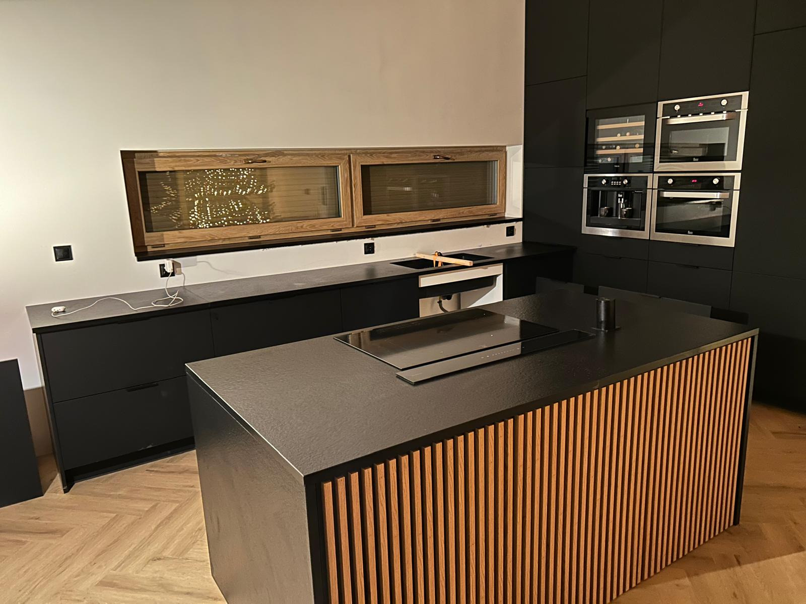 Nowoczesna kuchnia w ciemnej tonacji z wyspą wykończoną drewnianymi listwami i oknami z żaluzjami. Widoczne sprzęty AGD w zabudowie.