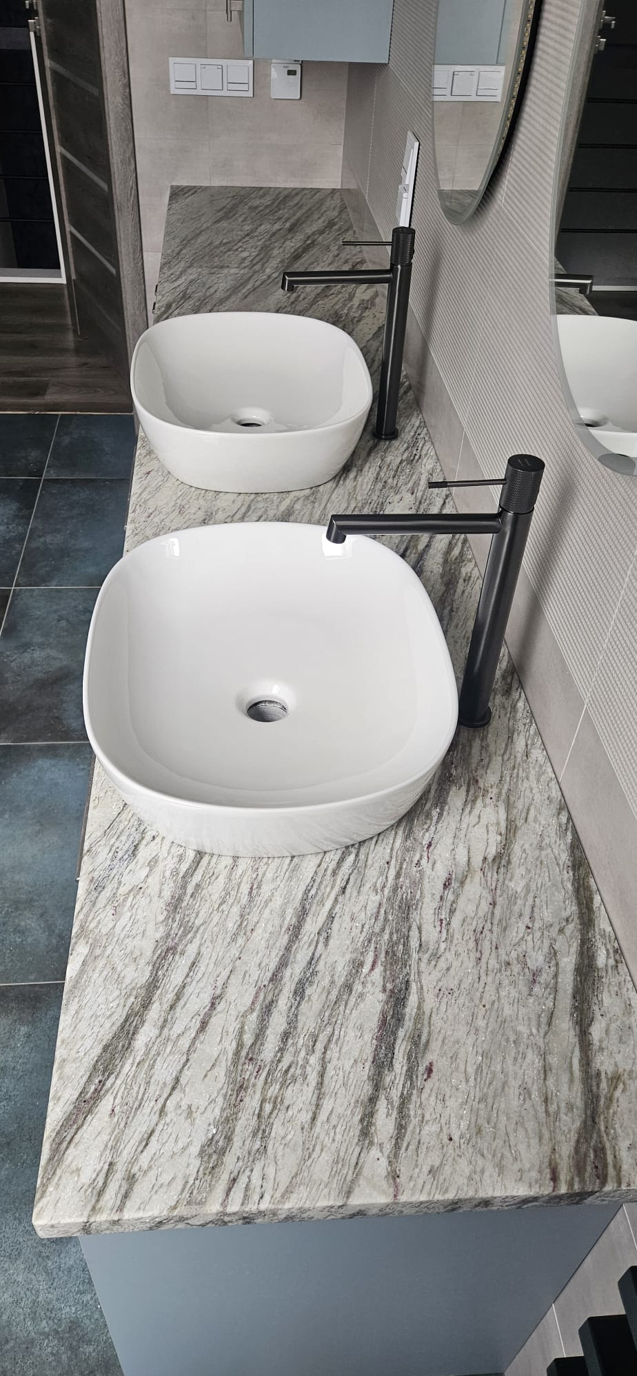 Dwie białe umywalki nablatowe o kwadratowym kształcie z zaokrąglonymi rogami, z czarnymi bateriami na blacie z wzorem przypominającym marmur.