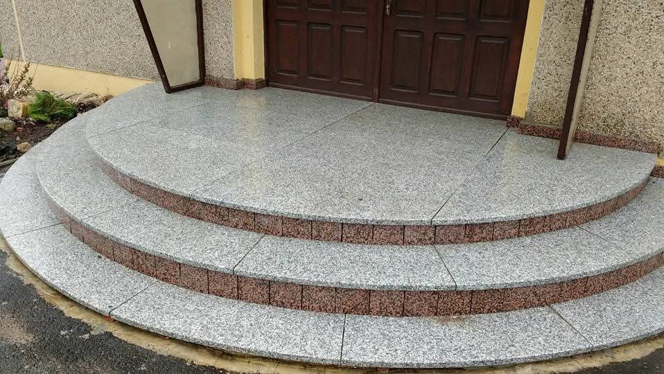 Granitowe schody wejściowe z ciemnymi obrzeżami prowadzące do brązowych drzwi. Elewacja budynku z szarą fakturą. Widok z dołu, na zewnątrz.