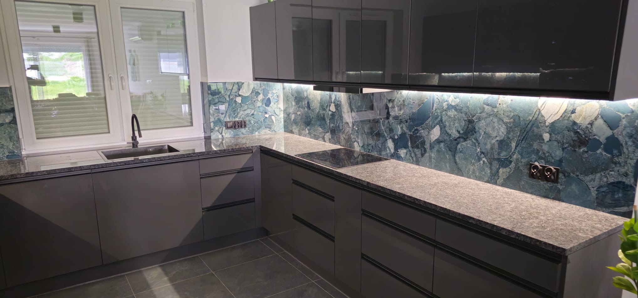 Nowoczesna kuchnia z szarymi szafkami, blatem z konglomeratu i oknem PCV. Ściana nad blatem wykończona dekoracyjną płytą imitującą kamień.