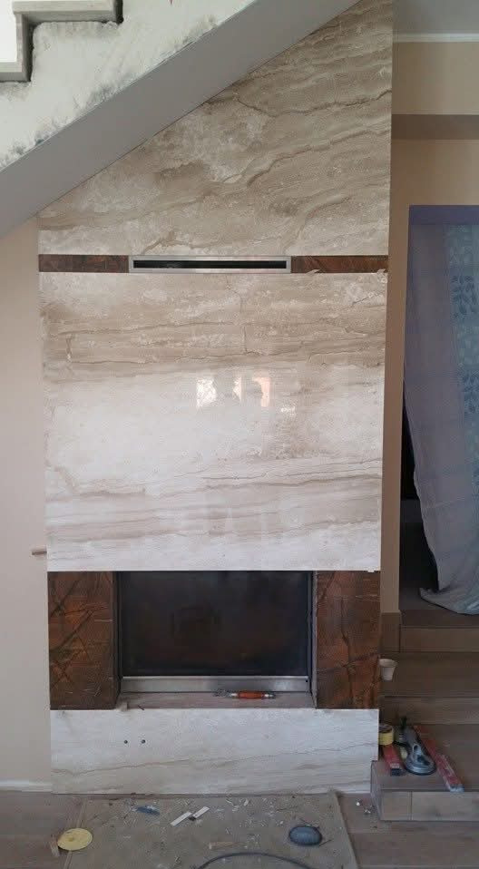 Obudowa kominka z beżowego marmuru z brązowymi akcentami, wkomponowana pod schody. Widoczne narzędzia i akcesoria montażowe na podłodze.