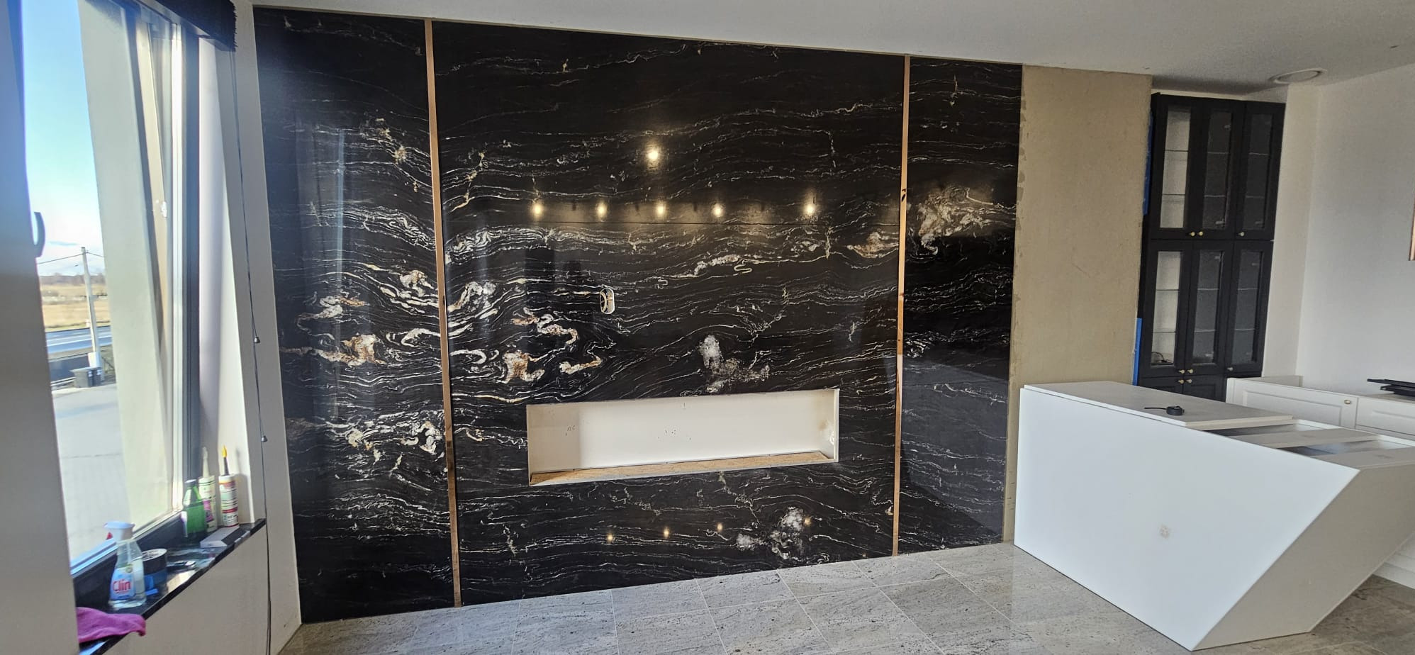 Czarny panel ścienny z marmurowym wzorem, oddzielony złotymi listwami, z wnęką na kominek, wnętrze w trakcie wykańczania.