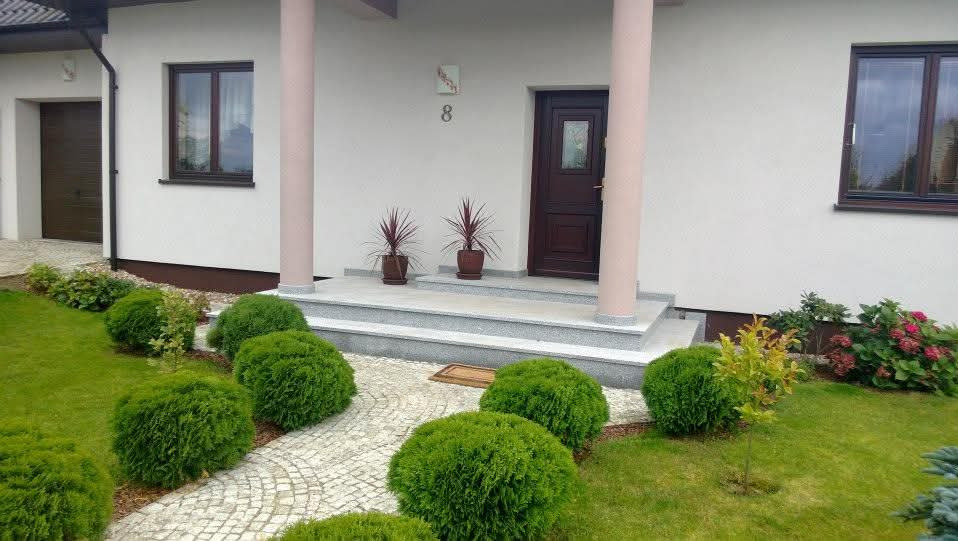 Elewacja domu z brązowymi oknami, drzwiami wejściowymi i kolumnami. Ogród z kulistymi krzewami i kamienną ścieżką prowadzącą do schodów.