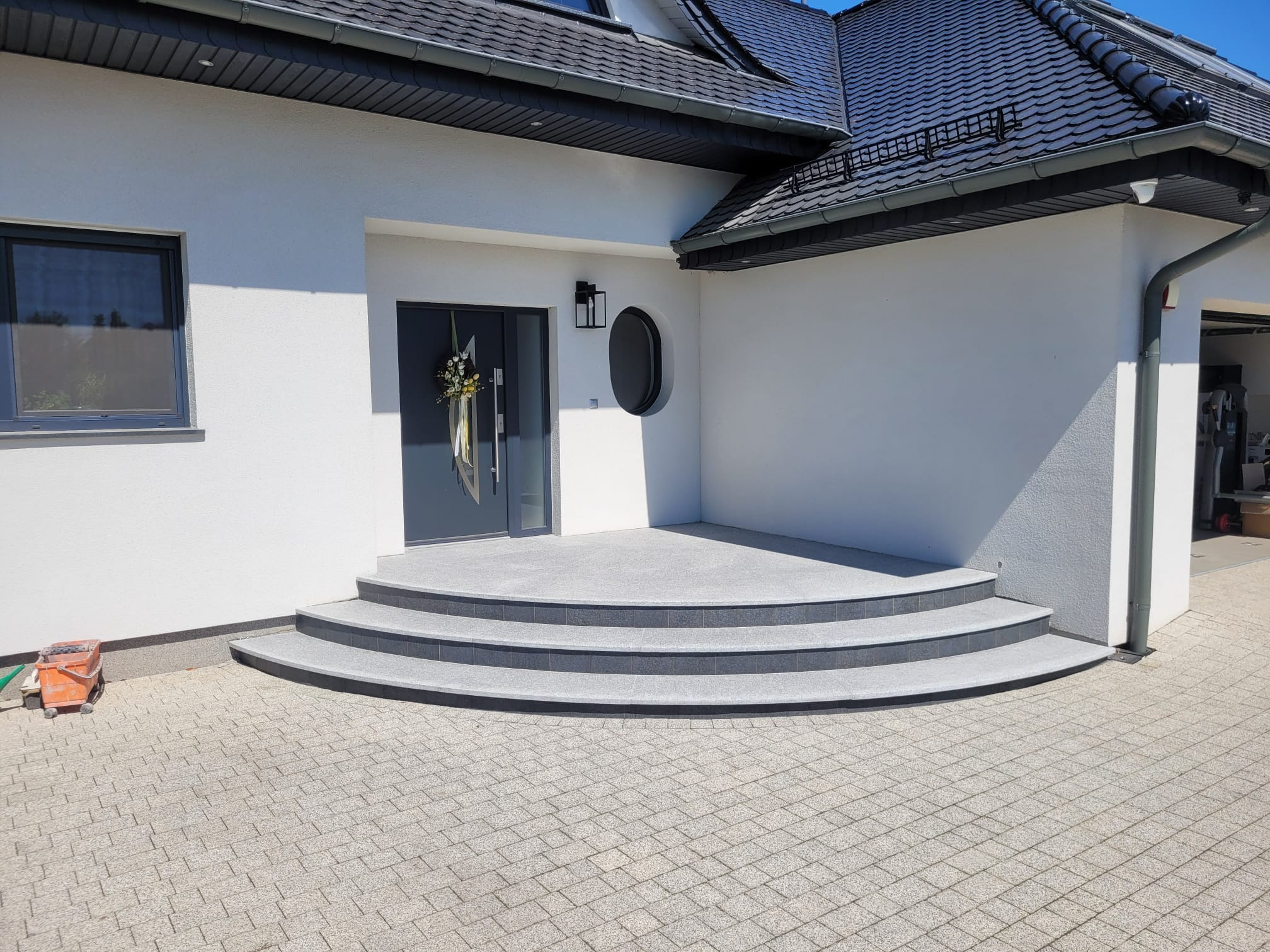 Eleganckie wejście do domu z szarymi schodami i drzwiami, nowoczesne oświetlenie zewnętrzne, jasna elewacja, kostka brukowa na podjeździe. Dekoracyjny wieniec na drzwiach.