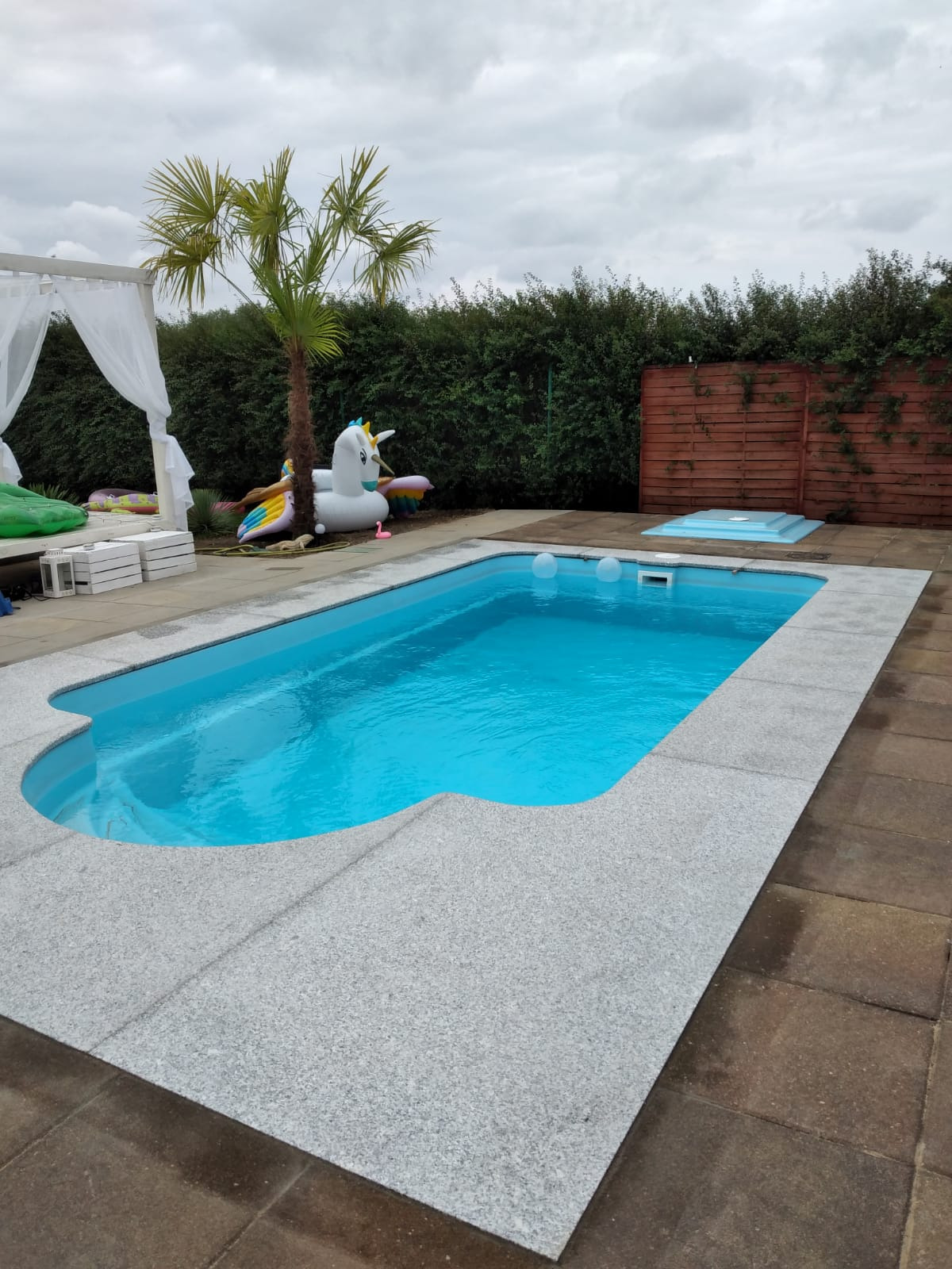 Błękitny basen z szarym wykończeniem, otoczony kostką brukową. W tle zielony żywopłot, drewniany płot, palma i dmuchany jednorożec przy białej altanie.