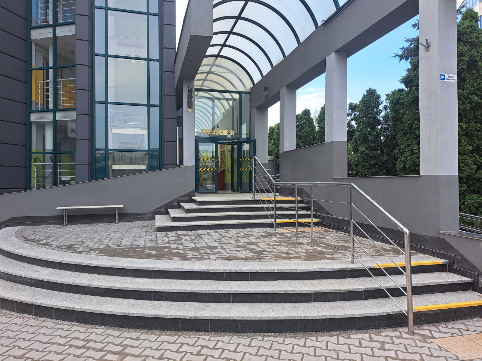 Nowoczesne wejście do budynku z przeszklonymi drzwiami oznaczonymi 'Wejście B', schodami z szarego granitu i metalową balustradą. Nad wejściem zadaszenie z przezroczystego tworzywa.