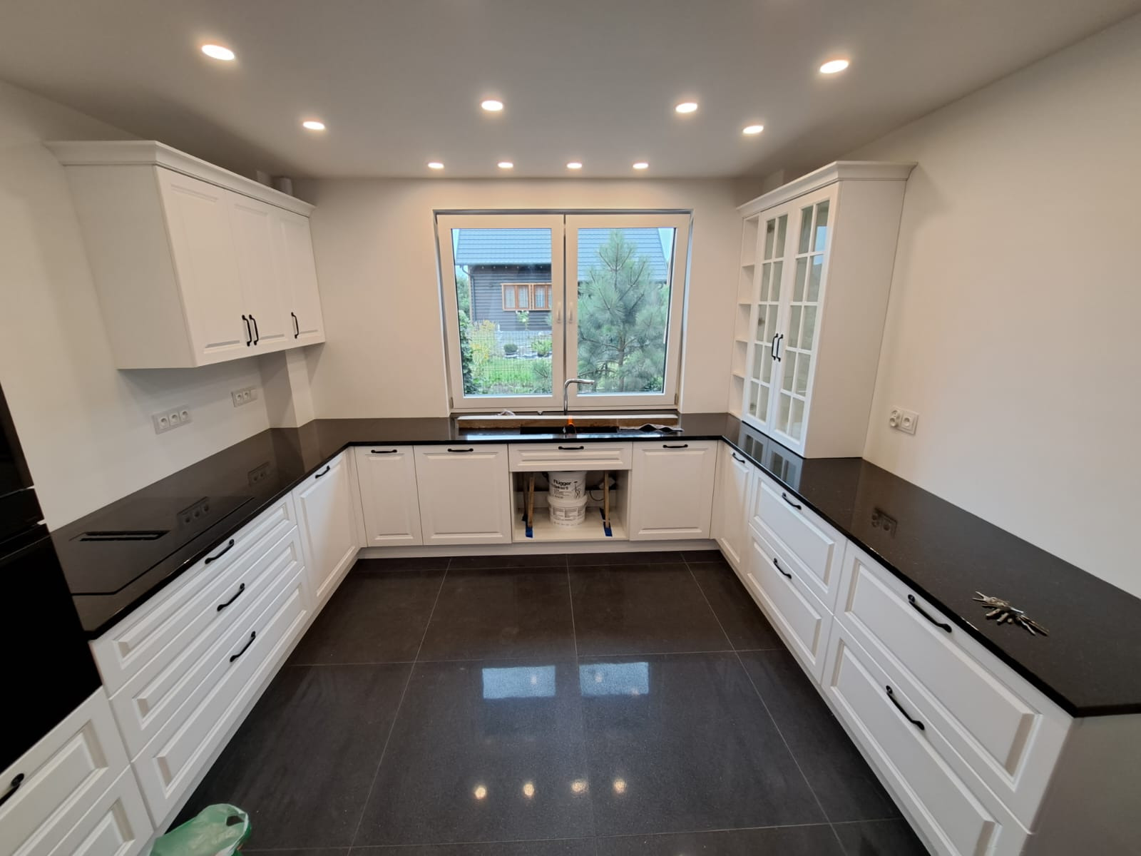 Nowoczesna kuchnia w bieli z czarnym blatem i dużym oknem, widok na ogród. Ustawienie w kształcie litery U, ciemna podłoga. Elementy wykończenia w stylu minimalistycznym.