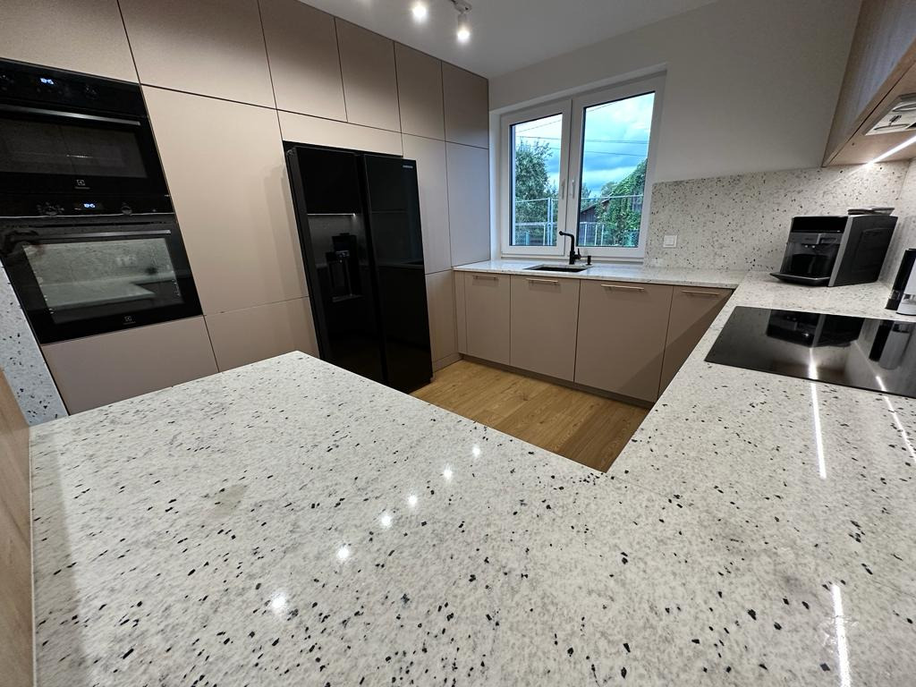 Kuchnia z beżowymi szafkami i blatem z konglomeratu w czarno-białe plamki. Widoczne okno, sprzęt AGD i drewniana podłoga.