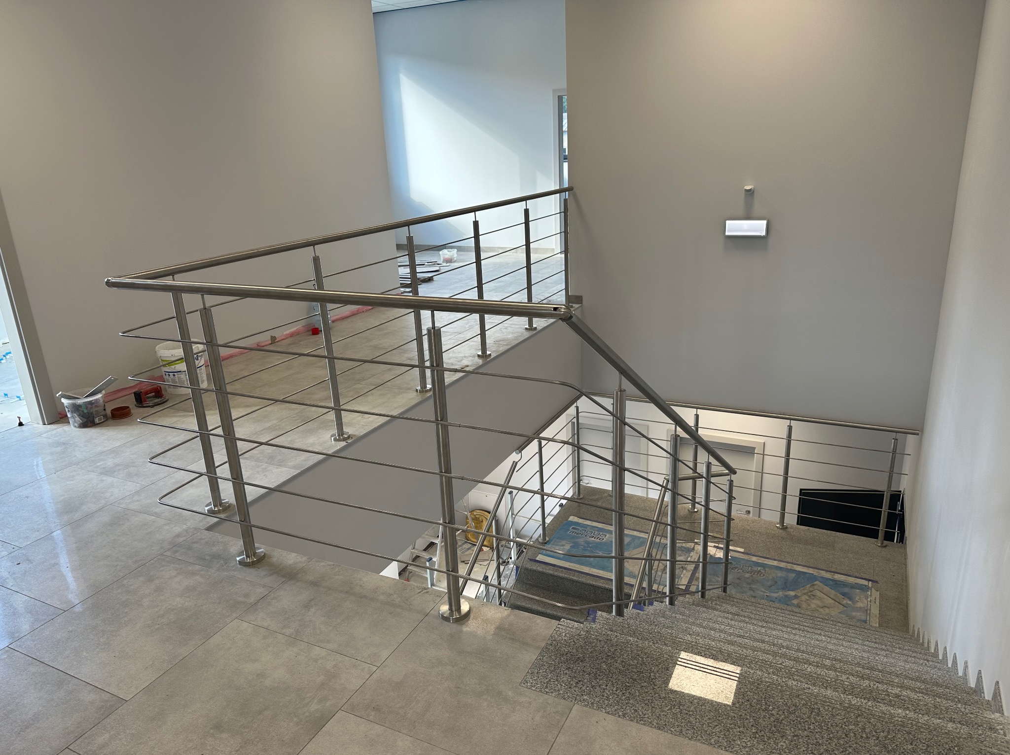 Nowoczesne, minimalistyczne balustrady ze stali nierdzewnej na schodach w Dąbrowie Górniczej. Widok z góry, jasne wnętrze, proste linie, szare płytki.