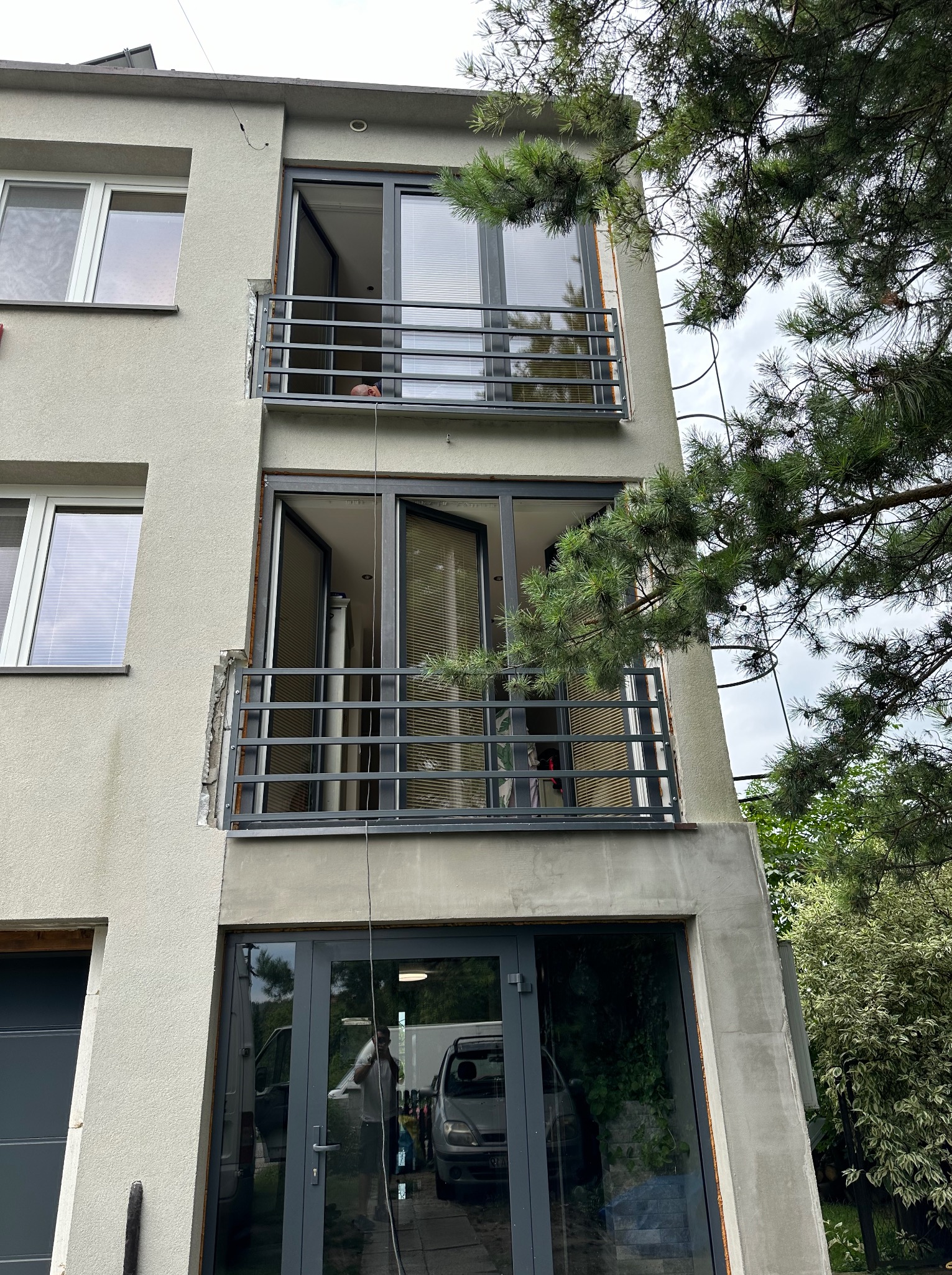 Szara fasada budynku z balkonami z metalowymi balustradami. Widoczne otwarte okna balkonowe i fragment drzewa iglastego po prawej stronie. U dołu okna z odbiciami.