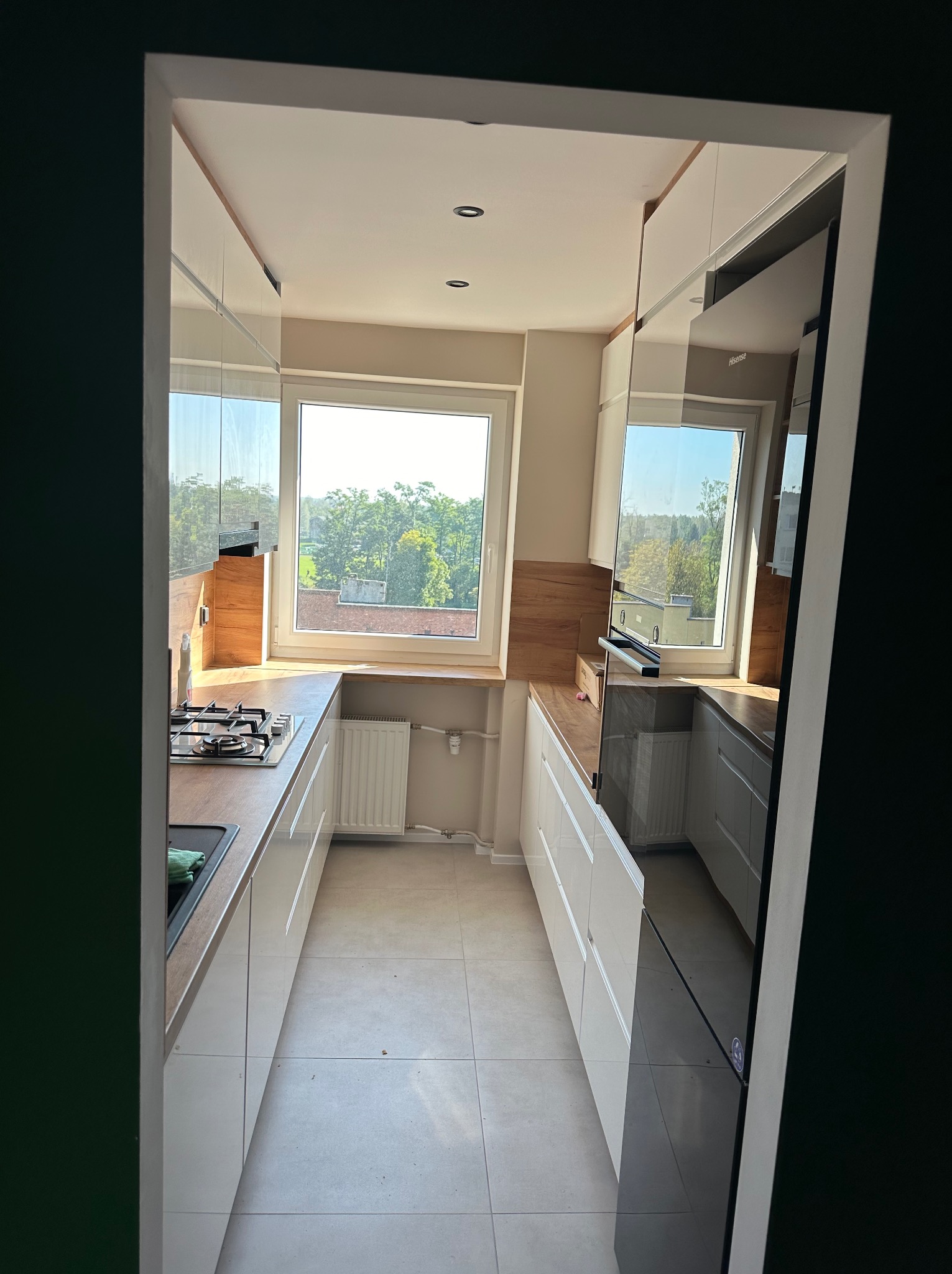 Nowoczesna kuchnia z białymi szafkami i drewnianymi blatami, widziana z perspektywy wejścia. Okno z widokiem na zieleń. Jasne płytki podłogowe.