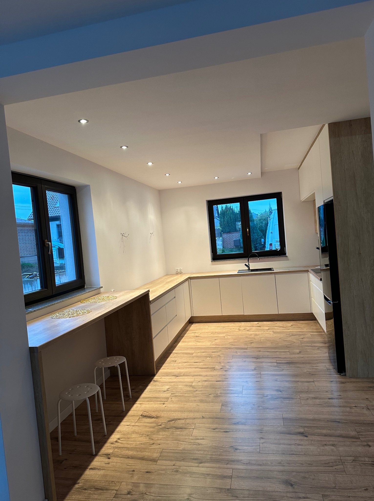 Jasna kuchnia z drewnianymi akcentami i dwoma oknami. Białe szafki, drewniany blat roboczy i podłoga. Dwa białe stołki barowe.