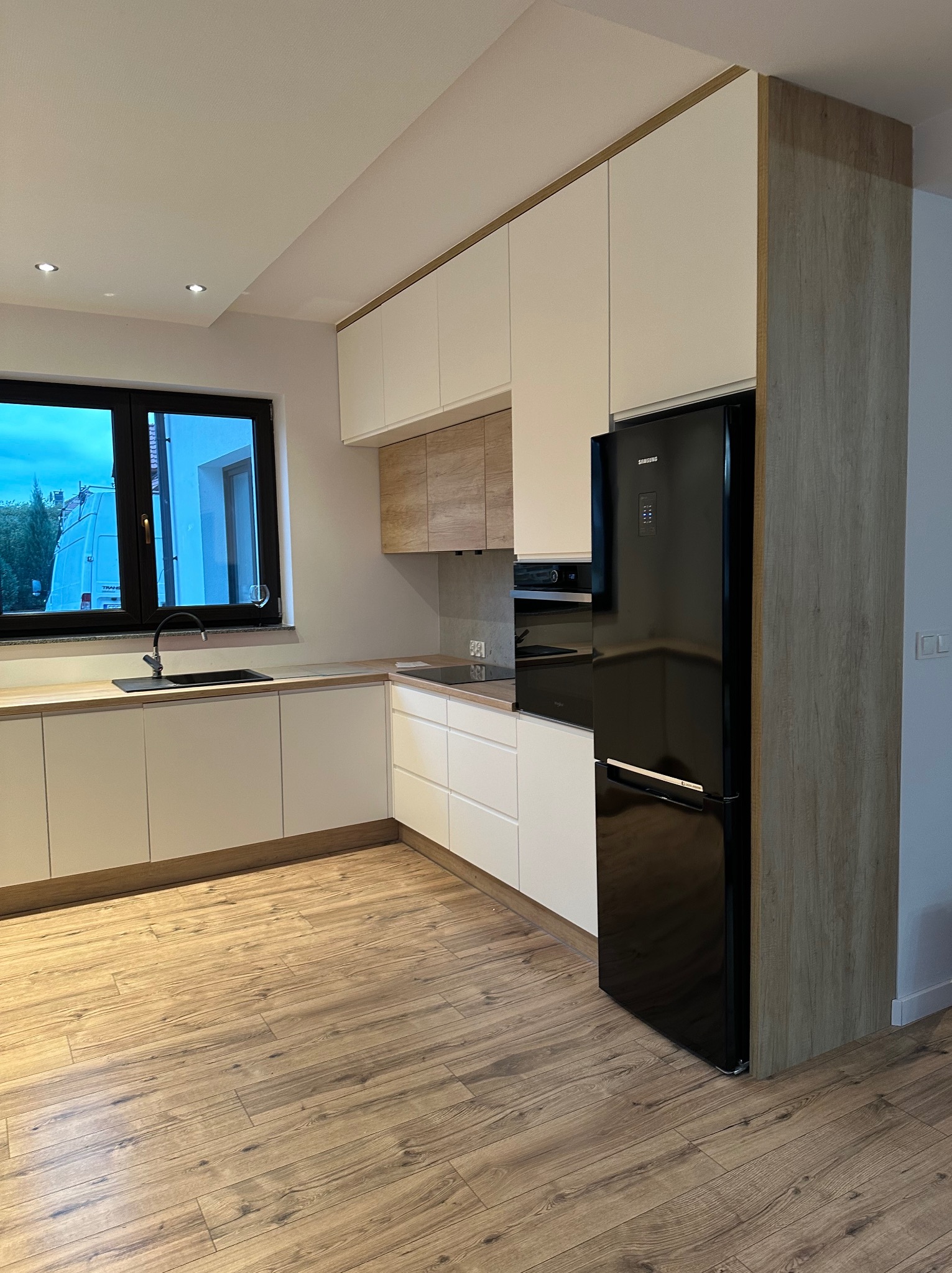Nowoczesna, jasna kuchnia na wymiar z białymi frontami, drewnianymi akcentami i czarną lodówką Samsung. Widok na okno z czarną ramą i drewniany blat.