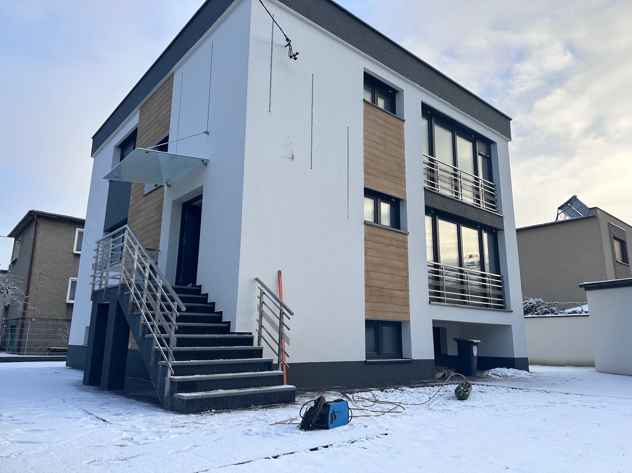 Nowoczesny dom z balkonami z metalową balustradą. Widoczne schody wejściowe z poręczą i sprzęt spawalniczy na śniegu. Dom ocieplony panelami imitującymi drewno.