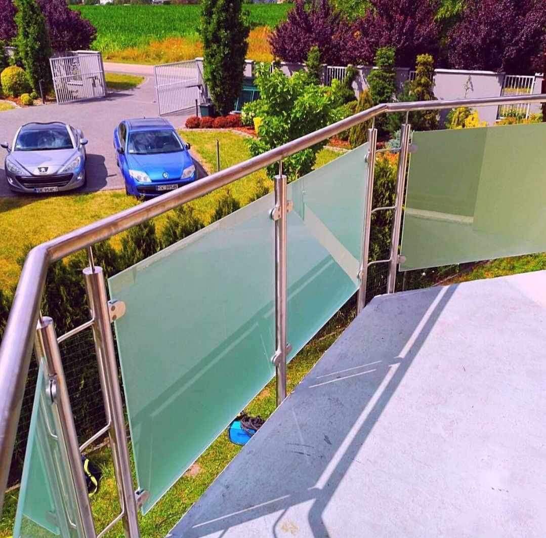 Szklana balustrada z nierdzewną poręczą na schodach zewnętrznych, w tle dwa samochody i brama wjazdowa.