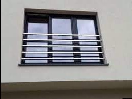 Nierdzewna, pozioma balustrada okienna z prostych rurek na tle jasnej elewacji. Czarne ramy okienne z trzema szybami. Minimalistyczny design.