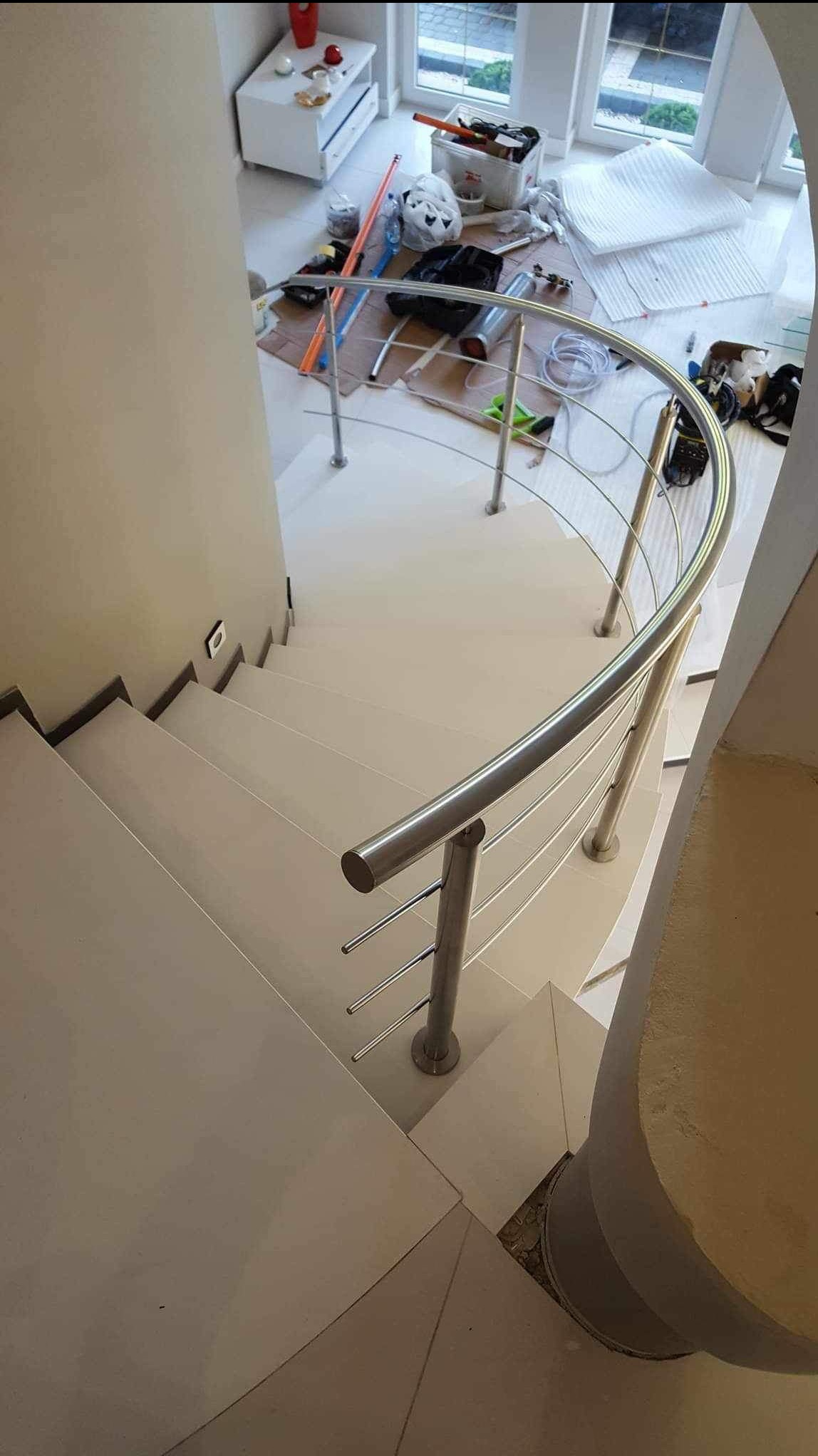 Montaż poręczy ze stali nierdzewnej na schodach w jasnym wnętrzu. Widok z góry ukazuje zakrzywiony kształt poręczy i białe stopnie. Częściowo widoczne narzędzia pracy.