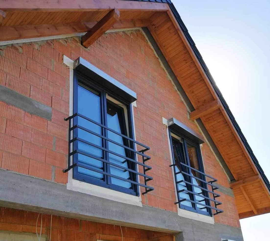 Dwa okna z czarnymi ramami i metalowymi balustradami na ceglanej ścianie domu pod skośnym dachem z drewnianą podbitką. Nowe budownictwo, surowy stan.