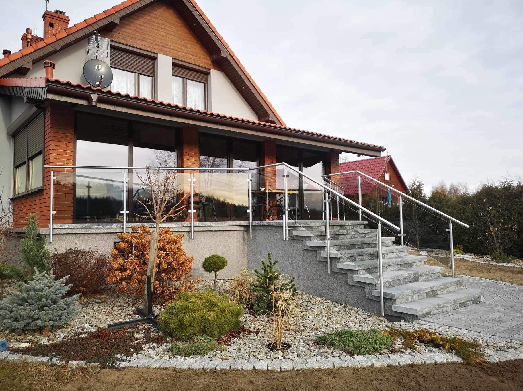 Nowoczesna szklana balustrada z metalowymi poręczami, otaczająca taras domu z cegły, z kamiennymi schodami i ozdobnym ogrodem na pierwszym planie.