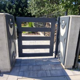 PMP-TECH - Nowoczesna, grafitowa furtka z geometrycznym wzorem, osadzona na betonowych słupkach. Widoczne detale mocowań i kostka brukowa. Tło stanowi zieleń ogrodu.