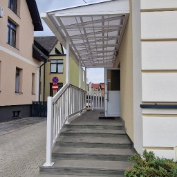 PMP-TECH - Zadaszone schody wejściowe z białą balustradą, prowadzące do budynku. Widoczne otoczenie budynku i fragment chodnika z kostki brukowej.