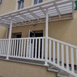 PMP-TECH - Biała balustrada z metalu i zadaszenie nad wejściem do budynku, widoczne schody i elementy konstrukcyjne. Ujęcie z boku budynku, zieleń w tle, fragment ulicy.