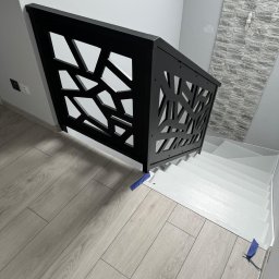 PMP-TECH - Nowoczesna, czarna balustrada schodowa z geometrycznym wzorem, zamontowana na białych schodach. Ściana z dekoracyjną okładziną w tle. Parkiet w odcieniach szarości.