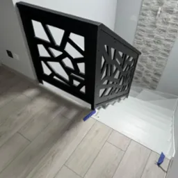 Nowoczesna, czarna balustrada schodowa z geometrycznym wzorem, zamontowana na białych schodach. Ściana z dekoracyjną okładziną w tle. Parkiet w odcieniach szarości.