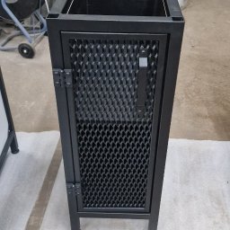 PMP-TECH - Czarna, metalowa szafka z perforowanymi drzwiczkami, widoczne zawiasy i uchwyt. W tle warsztat z elementami metalowymi. Nowoczesny design, surowy styl.