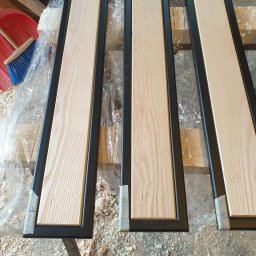 PMP-TECH - Elementy konstrukcyjne z drewna i metalu w warsztacie: trzy ramy z jasnym drewnem w czarnej metalowej obudowie, przygotowane do montażu, otoczone wiórami i narzędziami.