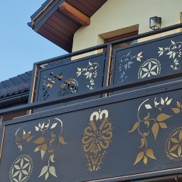 PMP-TECH - Czarna balustrada balkonowa z ażurowym wzorem roślinnym i geometrycznym, widoczna z dołu na tle jasnego budynku i błękitnego nieba w słoneczny dzień.