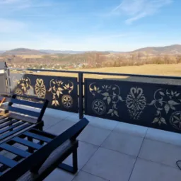 Czarna, metalowa balustrada balkonowa z ażurowym, złotym wzorem roślinnym, widok na g&oacute;rzysty krajobraz w tle. Na balkonie metalowe meble ogrodowe i narzędzia.