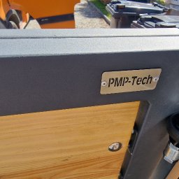 PMP-TECH - Fragment bramy przesuwnej z drewnianym wypełnieniem i tabliczką 'PMP-Tech', widoczny mechanizm jezdny z kółkiem, w tle pomarańczowy samochód dostawczy.