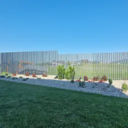 Nowoczesne ogrodzenie palisadowe z metalu, widok z ogrodu na tle błękitnego nieba. Elementy pionowe ogrodzenia tworzą rytmiczny wzór.