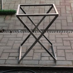 PMP-TECH - Metalowy stelaż stołu o surowym, minimalistycznym designie, wykonany z profili kwadratowych, z krzyżowym wzmocnieniem nóg. Stoi na brukowanej nawierzchni.