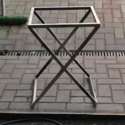 Metalowy stelaż stołu o surowym, minimalistycznym designie, wykonany z profili kwadratowych, z krzyżowym wzmocnieniem nóg. Stoi na brukowanej nawierzchni.