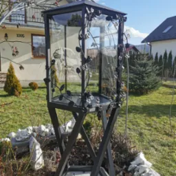 Kunsztowna, metalowa kapliczka z krzyżem i zdobieniami z motywem roślinnym, umieszczona w ogrodzie w Żywcu. Przeszklona konstrukcja na metalowej podstawie.