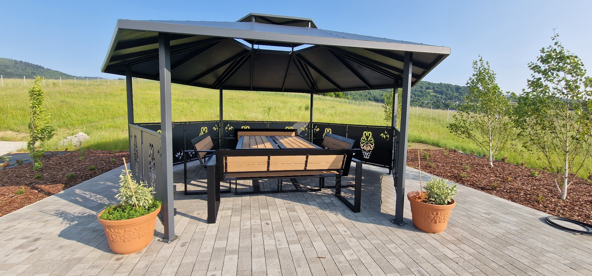 Metalowa pergola z ażurowymi panelami, stół z ławkami wewnątrz. Dwa donice z roślinami po bokach. Trawa i drzewa w tle, błękitne niebo. Całość w jasnych, naturalnych barwach.