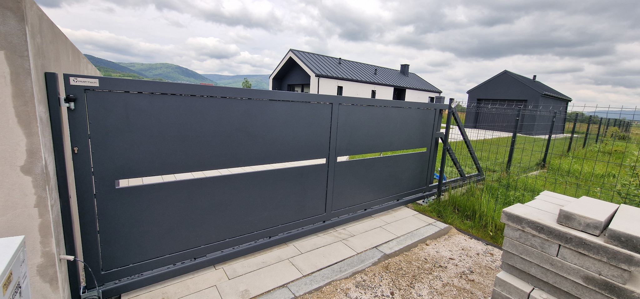 Nowoczesna, przesuwna brama wjazdowa w kolorze grafitowym z poziomymi wycięciami. W tle dom z ciemnym dachem i zielony krajobraz. Widoczne elementy konstrukcyjne i mechanizm bramy.