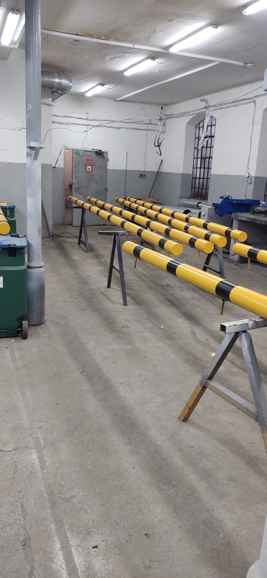 Żółto-czarne słupki na metalowych stojakach w warsztacie, widoczne okno i drzwi. Słupki pomalowane w pasy, równo ustawione w rzędach.