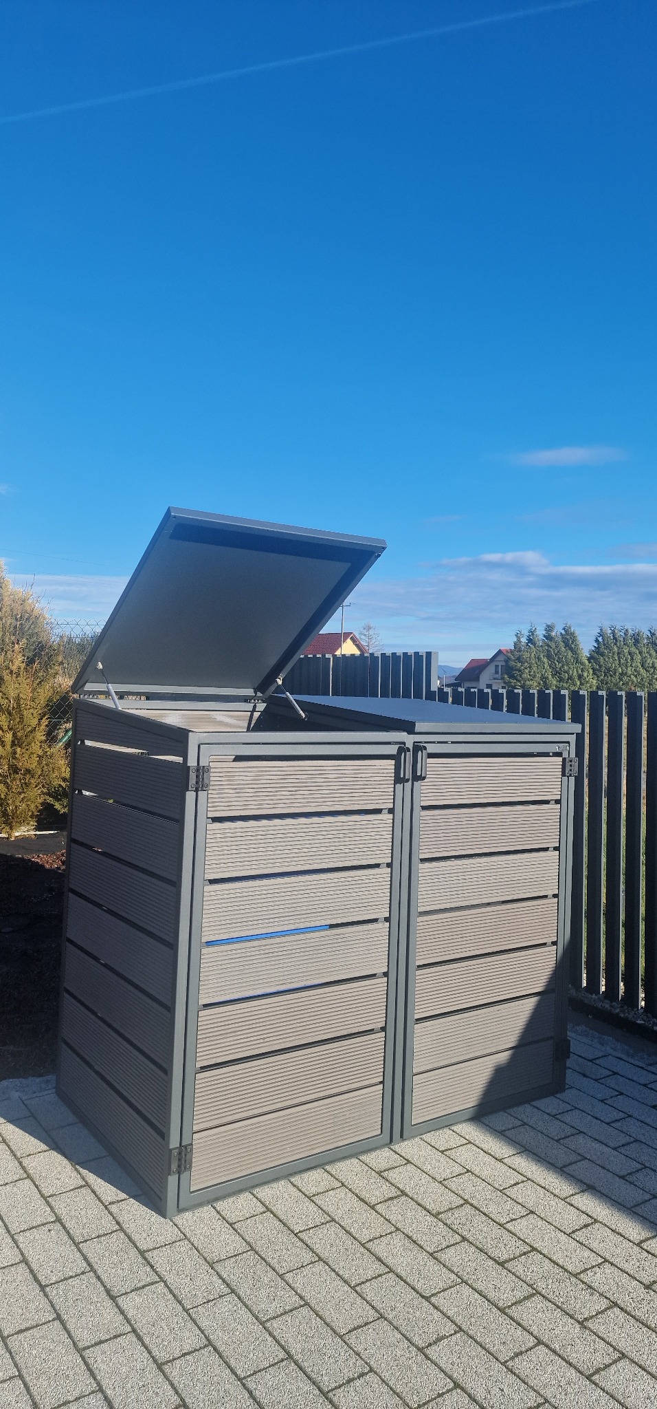 Metalowa zabudowa na dwa pojemniki na śmieci z podnoszoną klapą, z poziomych paneli w kolorze drewna, na tle kostki brukowej i ogrodzenia.