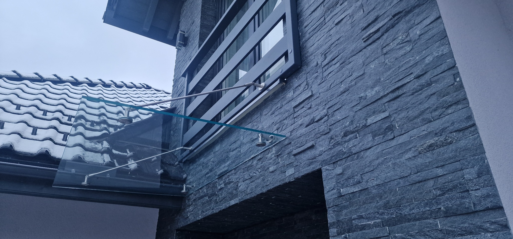 Szklany daszek nad wejściem, wsparty na metalowych elementach, z widoczną elewacją budynku wykończoną kamieniem łupkowym i fragmentem okna z metalową balustradą.