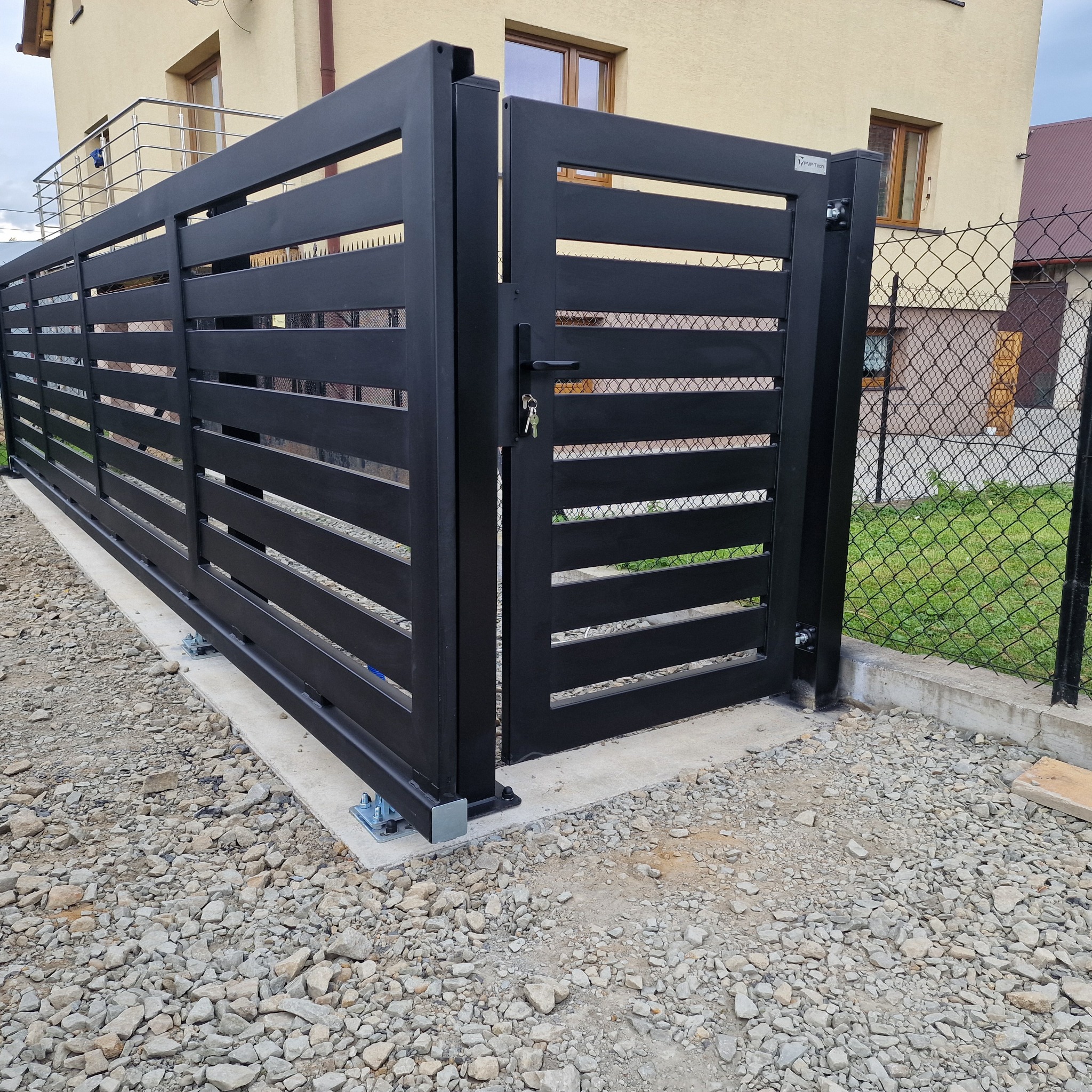 Nowoczesna, przesuwna brama wjazdowa z poziomymi panelami, pomalowana na czarno, z widocznym zamkiem i kluczem, osadzona na betonowej podstawie.