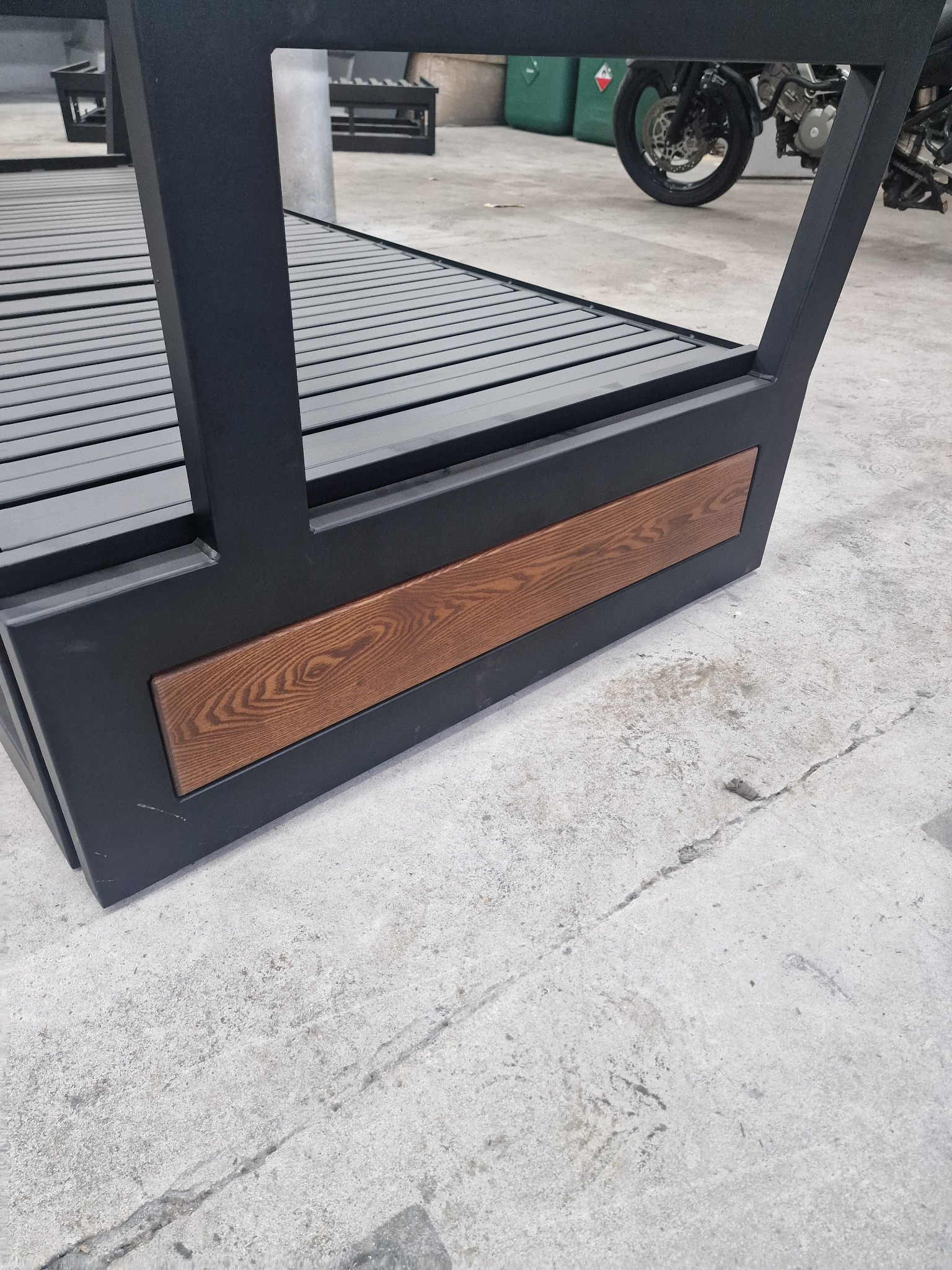 Czarno-szara konstrukcja metalowa z drewnianymi elementami dekoracyjnymi, przypominająca fragment pergoli, z motocyklem w tle. Widok z dołu, betonowa podłoga.
