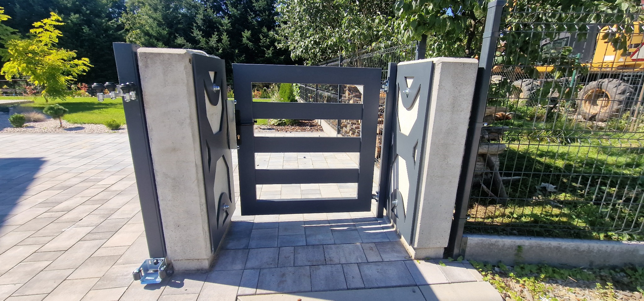 Nowoczesna, grafitowa furtka z geometrycznym wzorem, osadzona na betonowych słupkach. Widoczne detale mocowań i kostka brukowa. Tło stanowi zieleń ogrodu.