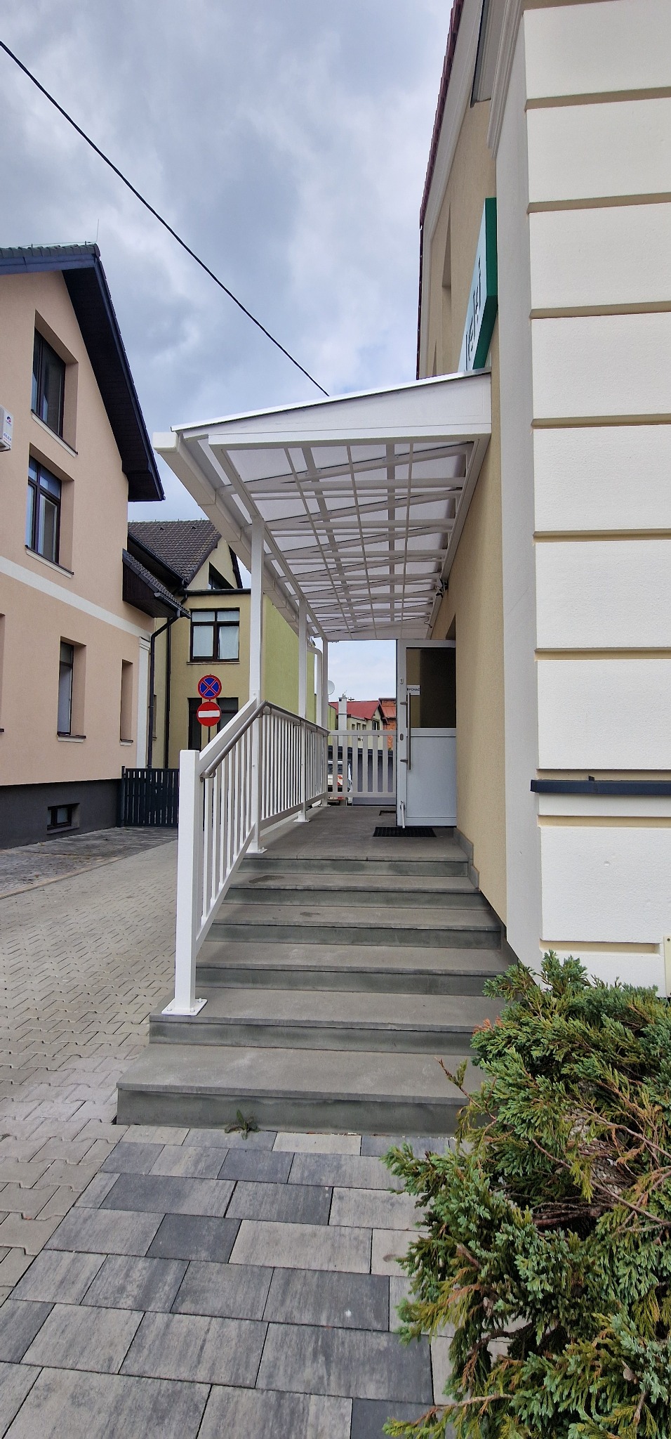 Zadaszone schody wejściowe z białą balustradą, prowadzące do budynku. Widoczne otoczenie budynku i fragment chodnika z kostki brukowej.