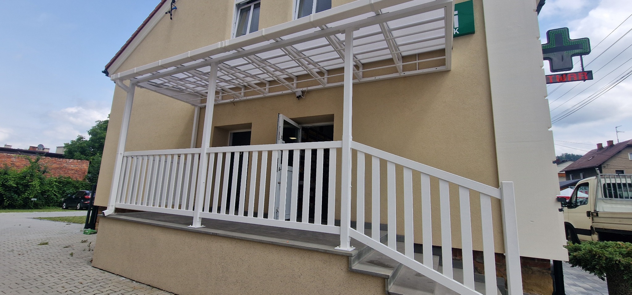 Biała balustrada z metalu i zadaszenie nad wejściem do budynku, widoczne schody i elementy konstrukcyjne. Ujęcie z boku budynku, zieleń w tle, fragment ulicy.