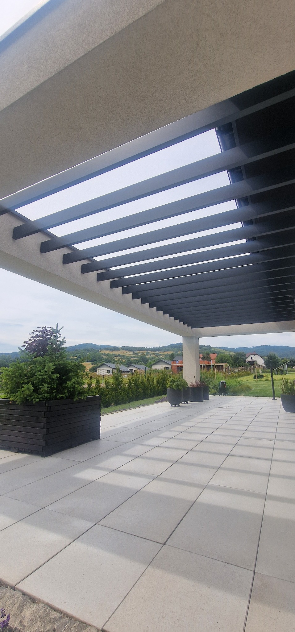 Nowoczesna pergola z szarymi, metalowymi lamelami i betonową konstrukcją, otoczona zielenią, z widokiem na wzgórza w tle. Minimalistyczny design, jasna przestrzeń.